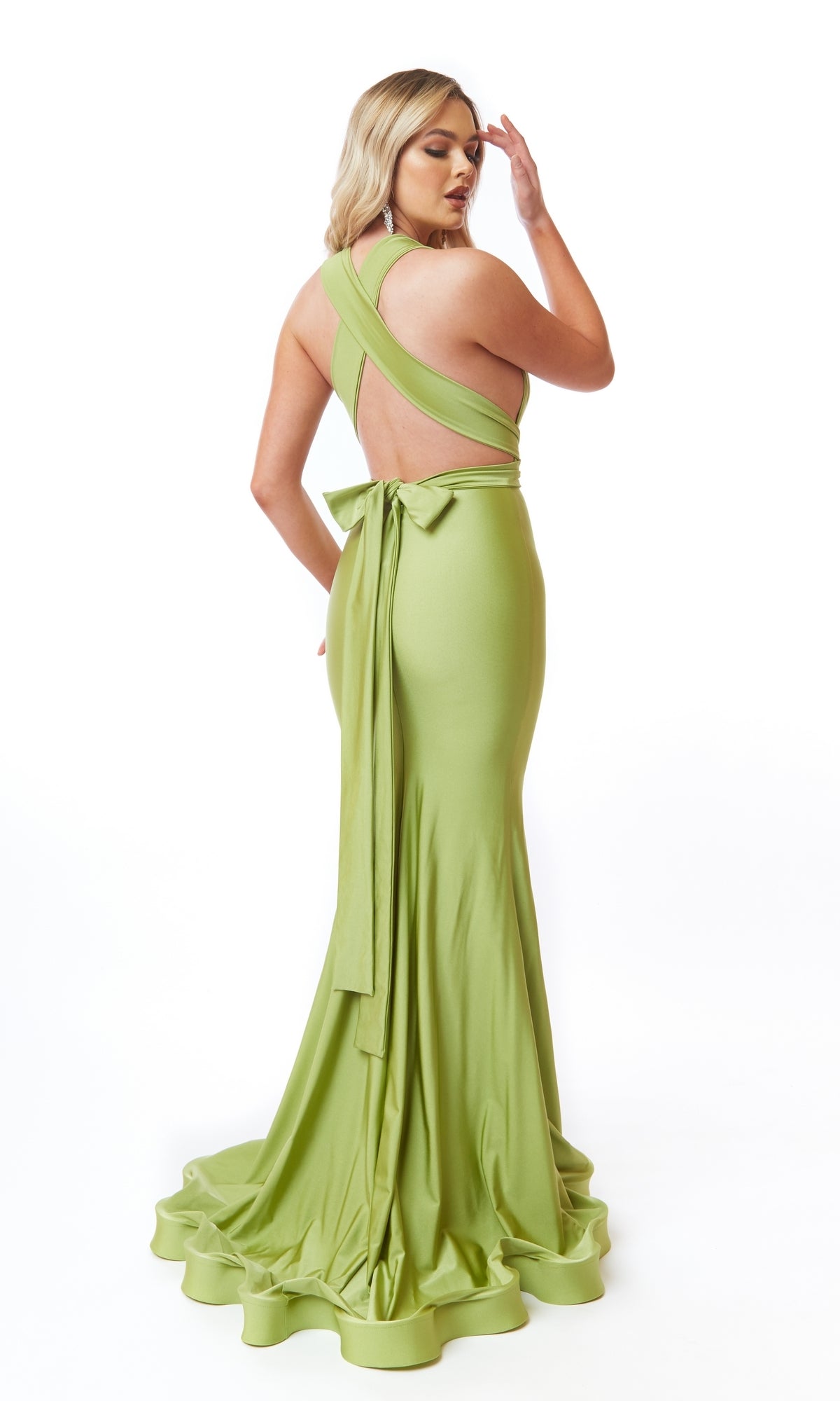 A woman models the Atria 6556H Back Bow Long V-Neck Prom Dress—a light green, floor-length, backless gown with crisscross straps, a mermaid silhouette, and a bow at the waist—against a white background.